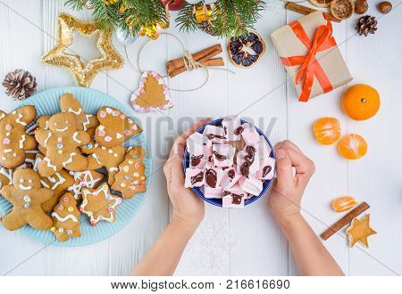 Top view female hands holding cup of hot cocoa with murshmellows on the wooden table with gift box, gingerbred cookies, christmas tree branches, decor, spices,tangerines. Selective focus. Text space.