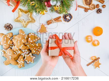 Top view female hands holding gift with red satin ribbon under white wooden table with gingerbred cookies, christmas tree branches with decor, spices and tangerines. Selective focus. Text space.