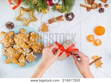 Top view female hands decorating gift with red satin ribbon on white wooden table with gingerbred cookies, christmas tree branches with decor, spices and tangerines. Selective focus. Text space.