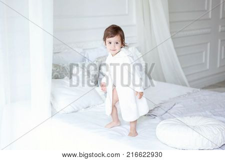 Funny and cute brunette little smiling girl playing on bed in bedroom. White interior with big bed