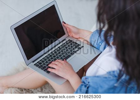 A young, beautiful bruneck woman in a denim cape holds a laptop in her lap sitting by the bed in front of a white background