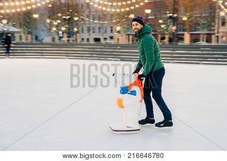 Outdoor Shot Of Glad Bearded Male Wears Green Jacket And Hat, Stands Near Skate Aid As Trying To Obt