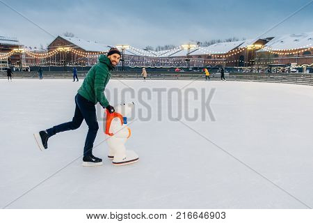 Outdoor Shot Of Glad Bearded Male Wears Green Jacket And Hat, Stands Near Skate Aid As Trying To Obt