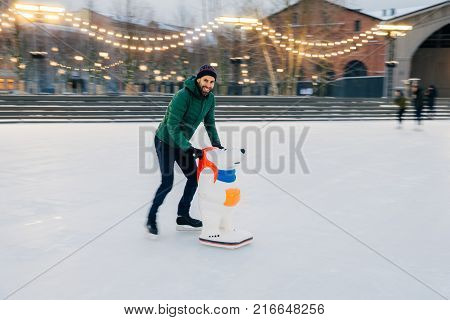 Outdoor Shot Of Unexperienced Male Skater Uses Skate Aid For Go Skating On Ice, Being In Good Mood,