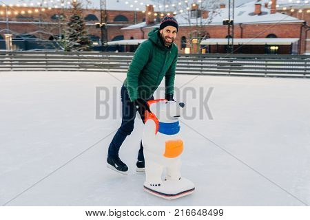 Outdoor Shot Of Unexperienced Male Skater Uses Skate Aid For Go Skating On Ice, Being In Good Mood,