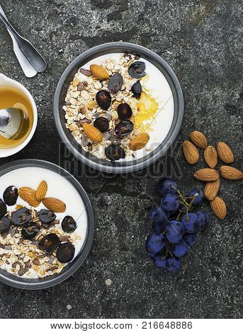 Breakfast from healthy seasonal ingredients: granola, flakes, honey, dark grapes, almonds in a serving gray pial on a gray grunge background. Top view. Vegetarian Organic Food Concept