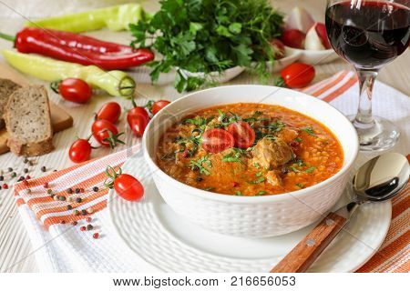 Traditional Georgian soup Kharcho with meat and rice in a white bowl next to bread, tomatoes, peppers, herbs, a glass of red wine on a white orange tablecloth. Horizontal. Close-up.