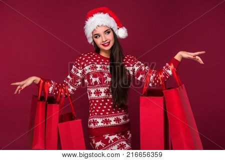 Young pretty woman or girl with long beautiful hair in Santa hat and New Year's dress holding red package on red studio background. Christmas sale. Emotional brunette. Copy space