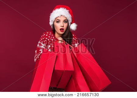 Young pretty woman or girl with long beautiful hair in Santa hat and New Year's dress holding red package on red studio background. Christmas sale. Emotional brunette. Copy space