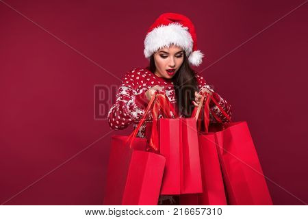 Young pretty woman or girl with long beautiful hair in Santa hat and New Year's dress holding red package on red studio background. Christmas sale. Emotional brunette. Copy space