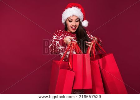 Young pretty woman or girl with long beautiful hair in Santa hat and New Year's dress holding red package on red studio background. Christmas sale. Emotional brunette. Copy space