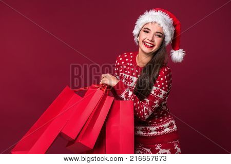 Young pretty woman or girl with long beautiful hair in Santa hat and New Year's dress holding red package on red studio background. Christmas sale. Emotional brunette. Copy space