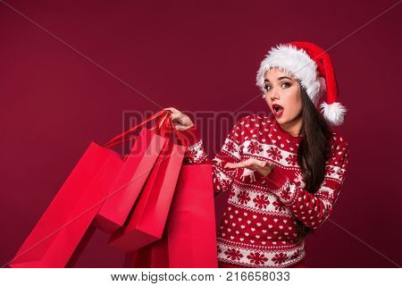 Young pretty woman or girl with long beautiful hair in Santa hat and New Year's dress holding red package on red studio background. Christmas sale. Emotional brunette. Copy space