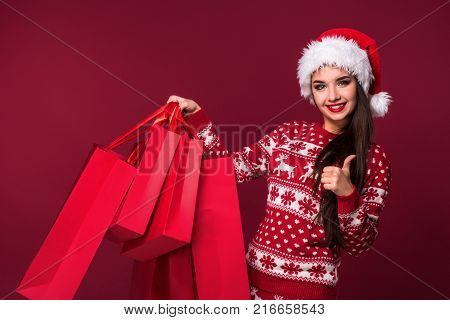 Young pretty woman or girl with long beautiful hair in Santa hat and New Year's dress holding red package on red studio background. Christmas sale. Emotional brunette. Copy space