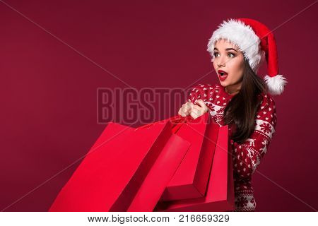 Young pretty woman or girl with long beautiful hair in Santa hat and New Year's dress holding red package on red studio background. Christmas sale. Emotional brunette. Copy space