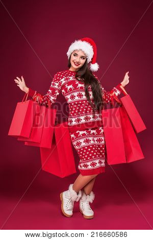 Young pretty woman or girl with long beautiful hair in Santa hat and New Year's dress holding red package on red studio background. Christmas sale. Emotional brunette. Copy space. Full-length photo