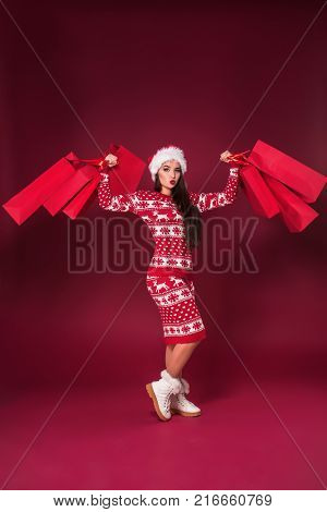 Young pretty woman or girl with long beautiful hair in Santa hat and New Year's dress holding red package on red studio background. Christmas sale. Emotional brunette. Copy space. Full-length photo