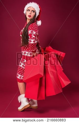 Young pretty woman or girl with long beautiful hair in Santa hat and New Year's dress holding red package on red studio background. Christmas sale. Emotional brunette. Copy space. Full-length photo