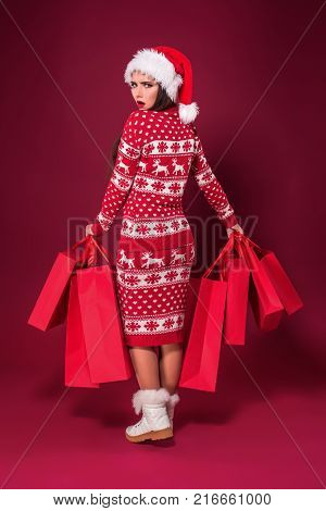 Young pretty woman or girl with long beautiful hair in Santa hat and New Year's dress holding red package on red studio background. Christmas sale. Emotional brunette. Copy space. Full-length photo