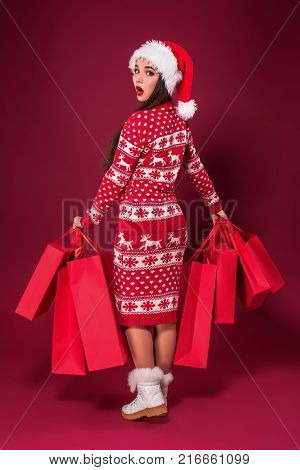 Young pretty woman or girl with long beautiful hair in Santa hat and New Year's dress holding red package on red studio background. Christmas sale. Emotional brunette. Copy space. Full-length photo