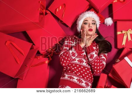 Funny young woman in a New Year's dress lying on a mountain of gifts. Christmas sale. Emotional brunette in Santa hat