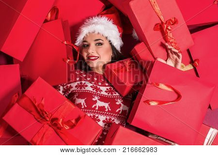 Funny young woman in a New Year's dress lying on a mountain of gifts. Christmas sale. Emotional brunette in Santa hat
