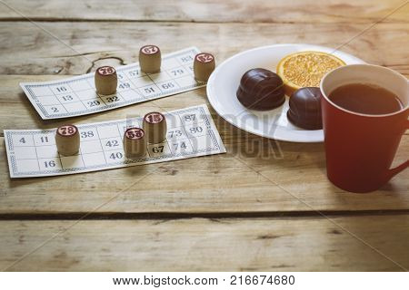 On A Wooden Bingo Table Game, A Cup With A Coffee, A Biscuit And An Orange Slice