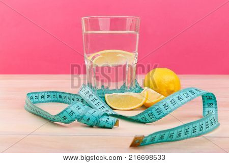 On the wooden table there is a glass with water and a slice of lemon, next to it there is a whole juicy lemon and a measuring tape. Side view. Inside.