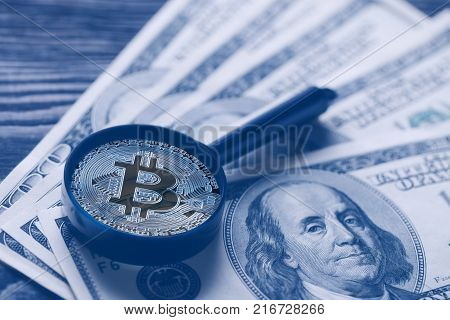 Bitcoin instead of magnifying glass over the background of hundred-dollar bills. Toning in blue. The concept of replacing paper money with virtual ones.