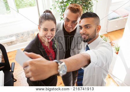Positive business friends taking self-portrait together. Excited purposeful young multiethnic managers posing for selfie on gadget in office. Colleagues concept
