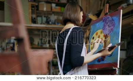 Talented young woman artist painting picture on canvas in art studio indoors