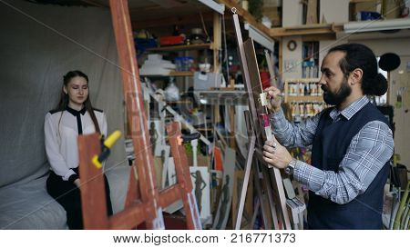 Sculptor creating sculpture of human's face on canvas while young woman posing him in art studio indoors