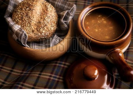 Food, a soup broth  and seeded Roll winter warming lunch, showing a rustic food scene with wooden bread dish and earthenware crockery soup dish with lid.