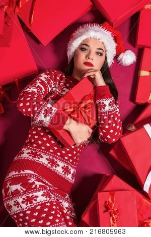 Funny young woman in a New Year's dress lying on a mountain of gifts. Christmas sale. Emotional brunette in Santa hat