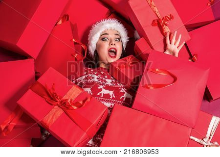 Funny young woman in a New Year's dress lying on a mountain of gifts. Christmas sale. Emotional brunette in Santa hat
