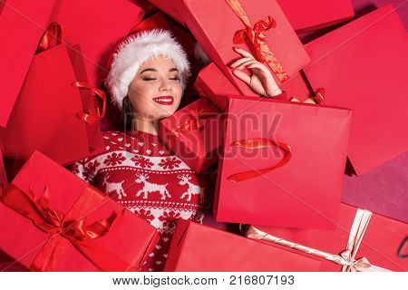 Funny young woman in a New Year's dress lying on a mountain of gifts. Christmas sale. Emotional brunette in Santa hat