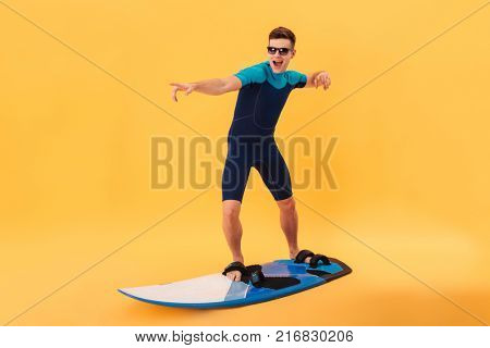 Image of happy surfer in wetsuit and sunglasses using surfboard like on wave over yellow background