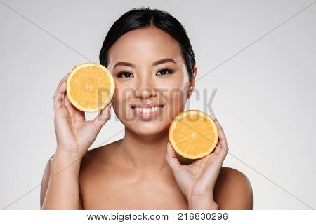Attractive half-naked asian woman amiling while holding orange slices near her face isolated over grey