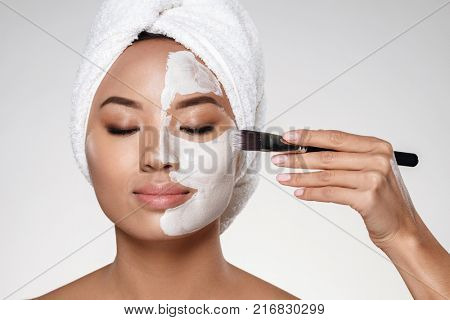 Young asian half-naked lady with towel on head put scrab on her face with brush isolated over grey