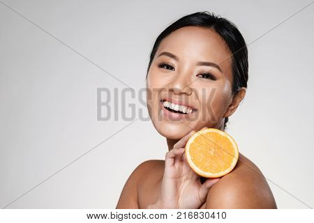 Attractive half-naked asian woman amiling while holding orange slices near her face isolated over grey
