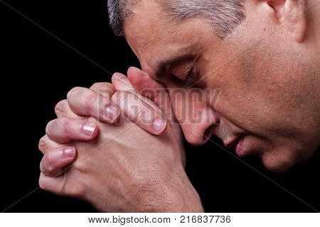 Close up of faithful mature man praying, hands folded in worship to god with head down and eyes closed in religious fervor. Black background. Concept for religion, faith, prayer and spirituality.