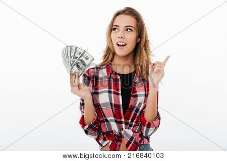 Portrait of an excited smiling girl in plaid shirt holding bunch of money banknotes and pointing finger up isolated over white background