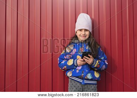 Happy girl kid with smartphone in hands on a red background in the street. Dressed in a blue jacket and smiling