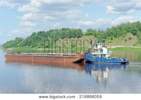 Volga, Russia. - June 2.2016. A river tug pushes a barge along the Volka River near a green bank covered with grass and bushes.