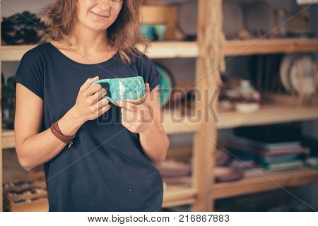 potter, tools, ceramics art concept - handsome young brunette female stand with finished utensils, smiling ceramist at workplace with baked cups and jugs at background