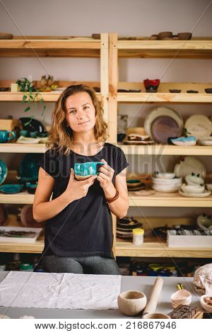 potter, tools, ceramics art concept - handsome young brunette female stand with finished utensils, smiling ceramist at workplace with baked cups and jugs at background