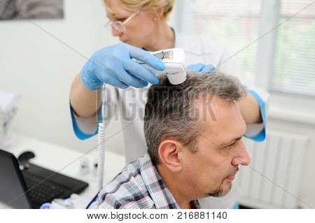 a man with gray hair at the reception at the dermatologist. the doctor examines the patient's hair using a method trihoskopii