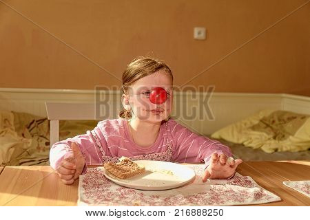 Kid girl playlful eats chocolate cream spread on bread. Chocolate sweet food snack. Happy girl has a snack in the kitchen. A cute small girl smiles. A small girl with chocolate cream stains on face. Little girl wears clown nose. Childhood and fun concept
