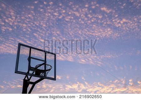 Silhouette of outdoor basketball goal with clear backboard and sunset in the background visible through backboard