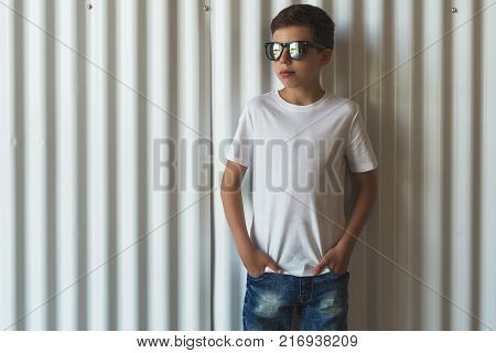 Front view. Young hipster boy in sunglasses dressed in white t-shirt is stands indoor against white wall. Mock up. Space for logo, text, image.
